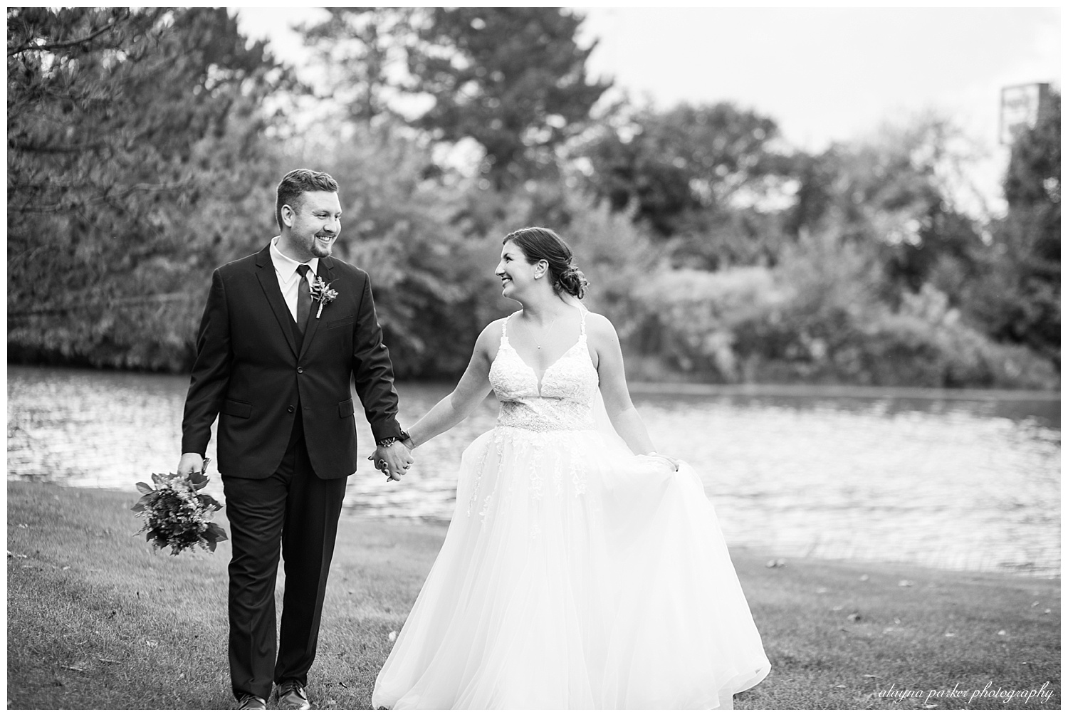 black and white image of bride and groom walking beside pond at water's edge
