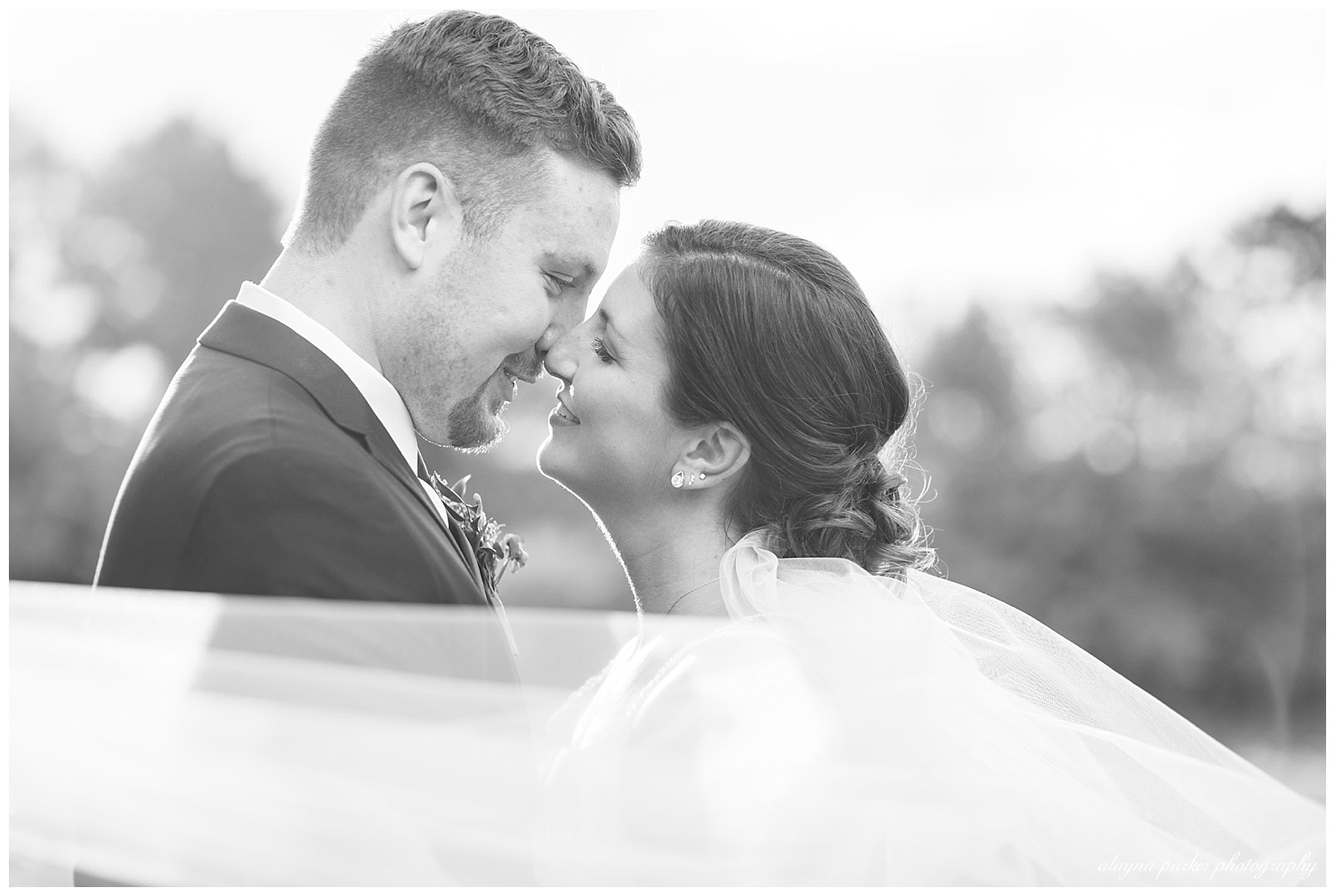 black and white image of bride and groom almost kissing while veil blows in wind at water's edge