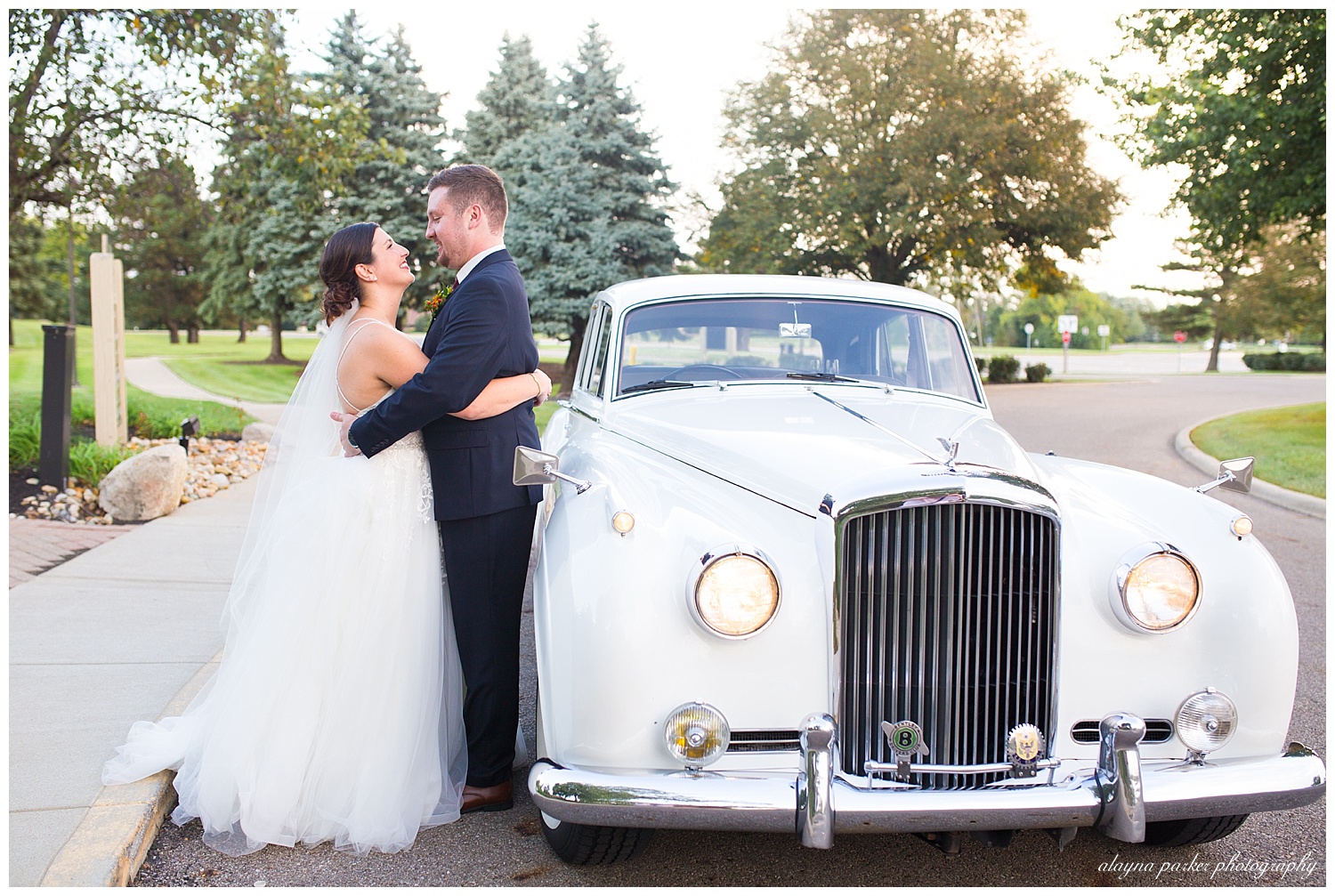 bride and groom hugging beside a white 1960's classic car at water's edge