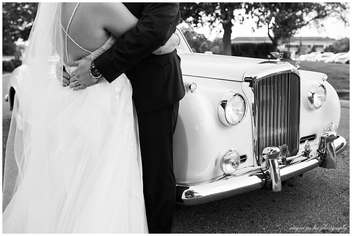 black and white image of close up of bride and groom hugging beside a white 1960's classic car at water's edge