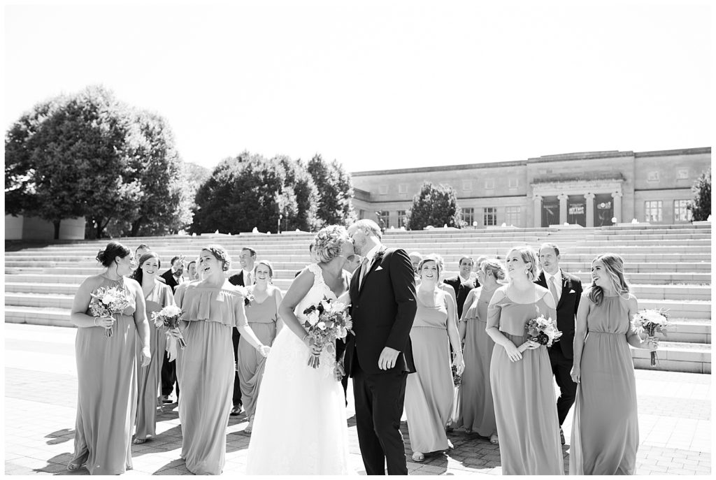 black and white image of bride and groom kissing while bridal party is in background in front of steps at cosi