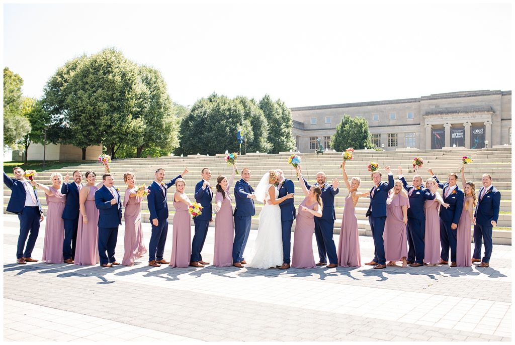 bridal party cheering while bride and groom kiss in front of steps at cosi