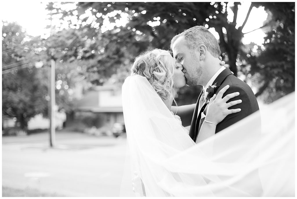 black and white image of bride and groom kissing while veil blows in wind