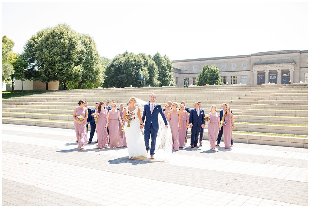 bride and groom walking while bridal party walks behind them in front of steps at cosi
