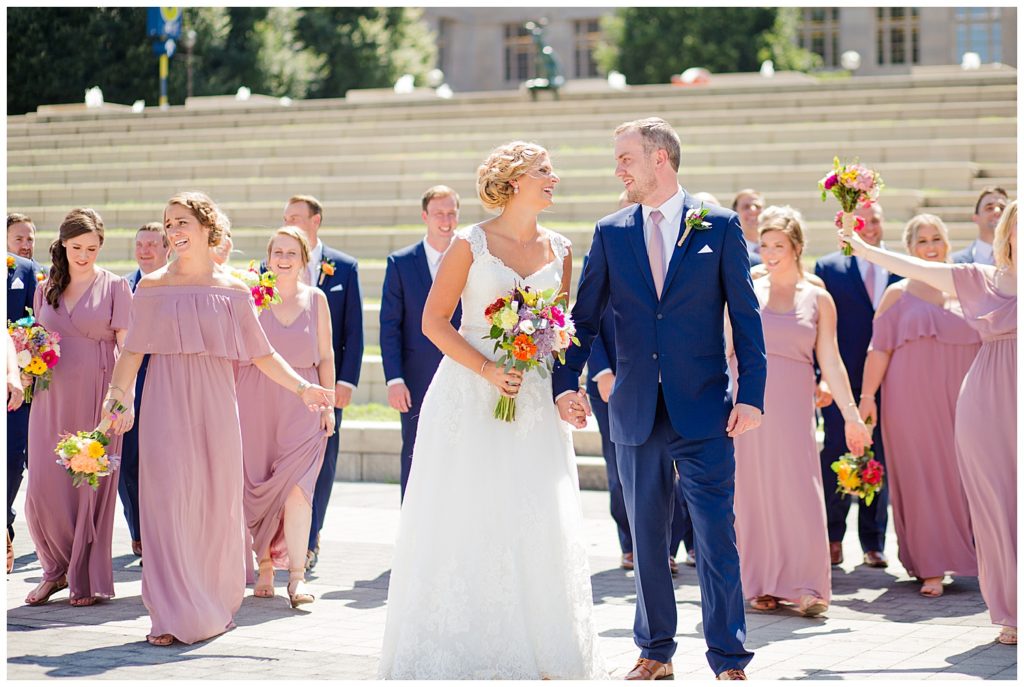 bride and groom laughing at each other while bridal party walks in background in front of steps at cosi