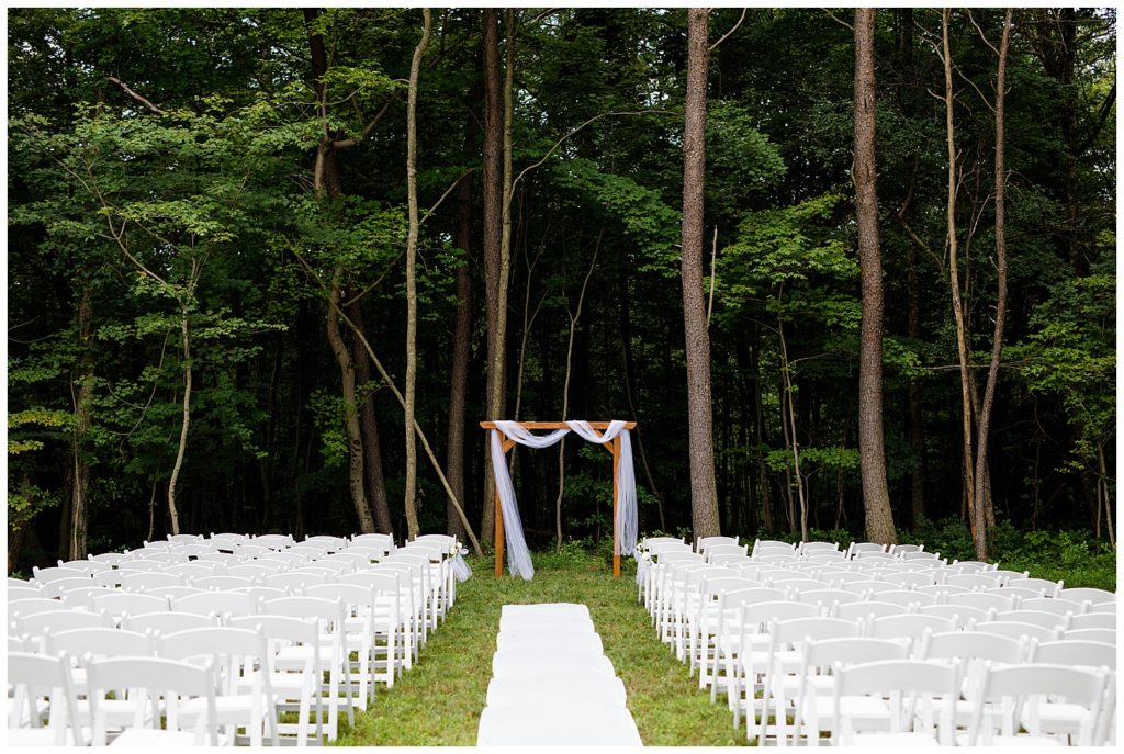 An image of a rustic outdoor wedding arch draped in a chiffon scarf with a background of trees with white chairs set up for guests at the Cedar Grove Lodge wedding venue by Columbus Ohio wedding photographer, Alayna Parker Photography