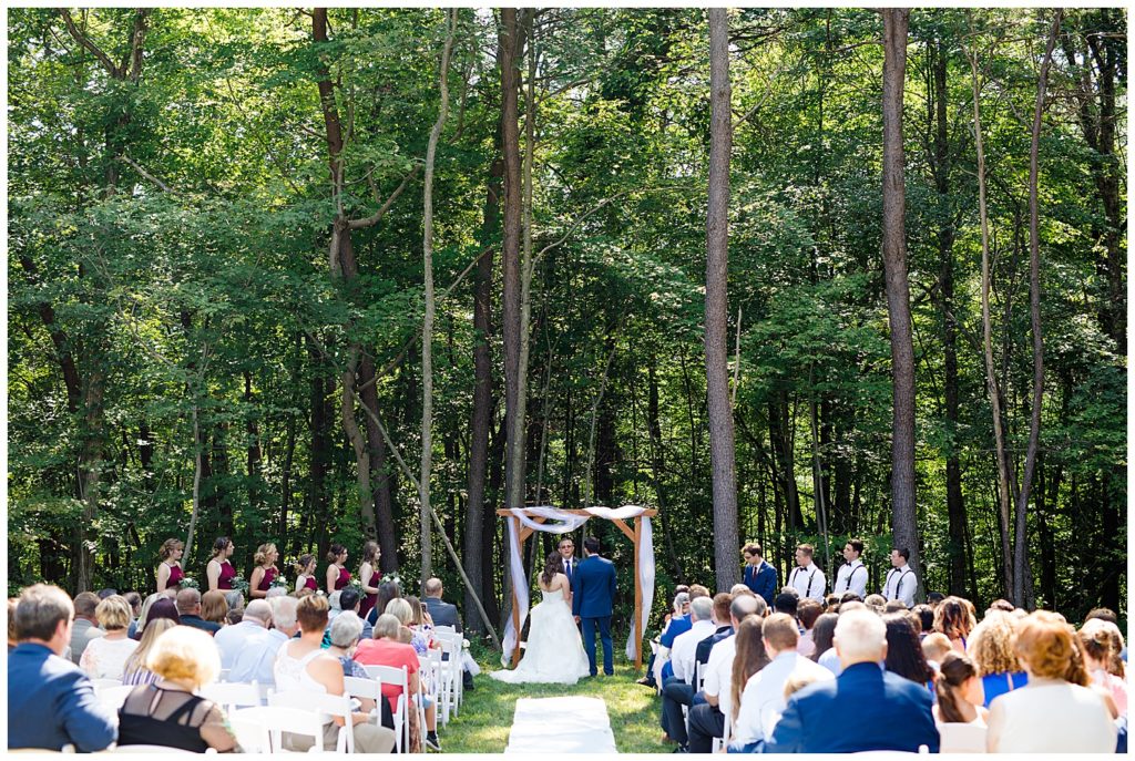 An image of a long-range view of the bride and groom standing with the wedding party during the wedding ceremony in a rustic outdoor setting at the Cedar Grove Lodge wedding venue by Columbus Ohio wedding photographer, Alayna Parker Photography
