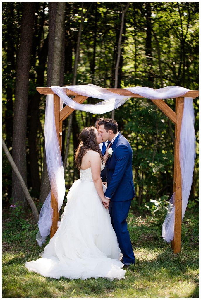 An image of the bride and groom kissing at the end of the wedding ceremony, framed by a rustic arch and trees at the Cedar Grove Lodge venue in Hocking Hills, Ohio by Columbus Ohio wedding photographer, Alayna Parker Photography