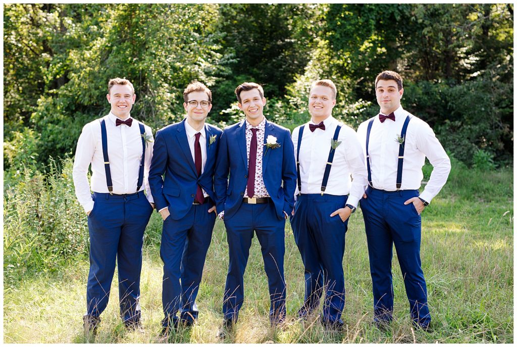 groomsmen wearing white shirt, blue suit pants, and suspenders and smiling at camera