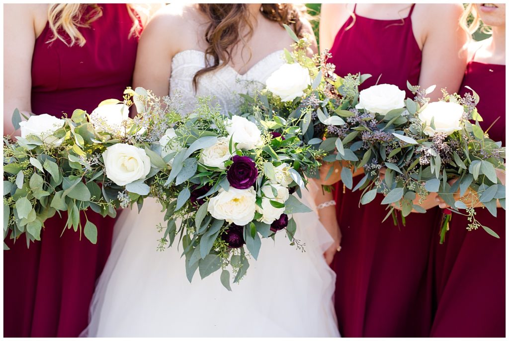 close up of bride and bridesmaids holding bouquets of eucalyptus, white flowers, and dark purple flowers