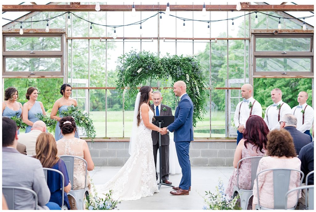 A photograph of the bride and groom holding hands as the officiant repeats the vows at their wedding ceremony at an Jorgensen farms Oak Grove wedding venue by Columbus OH wedding photographer, Alayna Parker Photography