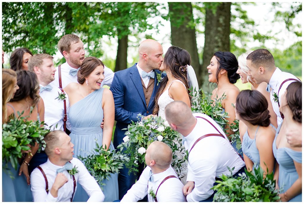 A picture of the bride and groom kissing as the wedding party relaxes and cheers them on after the wedding ceremony at an Jorgensen farms Oak Grove wedding venue by Columbus wedding photographer, Alayna Parker Photography