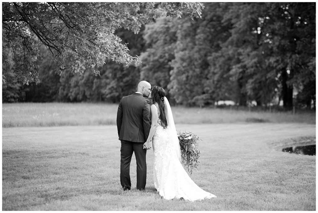 A photograph in black and white of the bride and groom pausing to kiss as they walk outdoors hand and hand at the Oak Grove venue in New Albany, Ohio by Columbus OH wedding photographer, Alayna Parker Photography