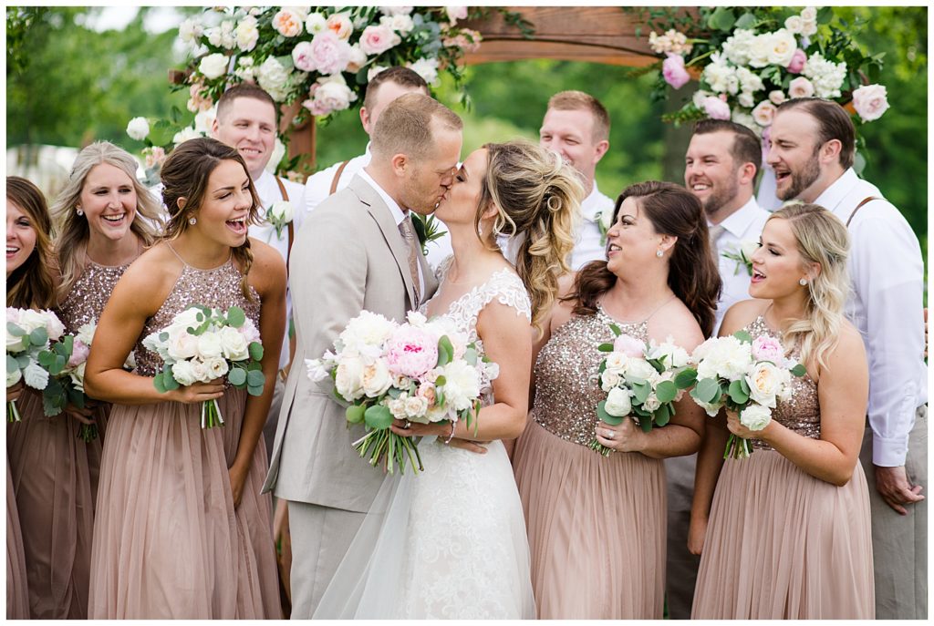 bridal party laughing while bride and groom kiss