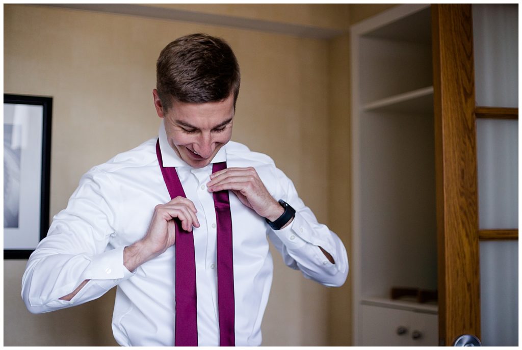 An image of a groom smiling and putting on his tie as he gets dressed for his wedding by Columbus Ohio wedding photographer, Alayna Parker Photography