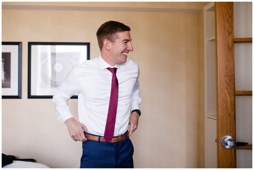 groom smiling at groomsmen while he gets dressed at Renaissance Hotel by Columbus wedding photographer, Alayna ParkerA picture of a groom smiling and adjusting his belt as he dresses for his wedding by Columbus wedding photographer, Alayna Parker Photography