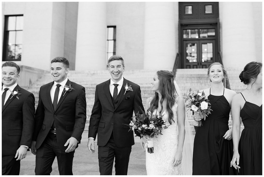 black and white image of groom smiling at camera while walking with bridal party outside ohio statehouse