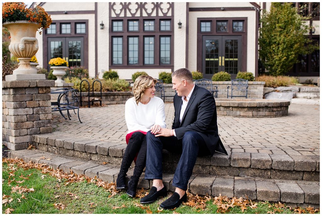 An image of an engaged couple holding hands, sitting outside on brick steps, smiling and gazing at each other at Brookside Golf & Country Club by Columbus Ohio engagement photographer, Alayna Parker Photography