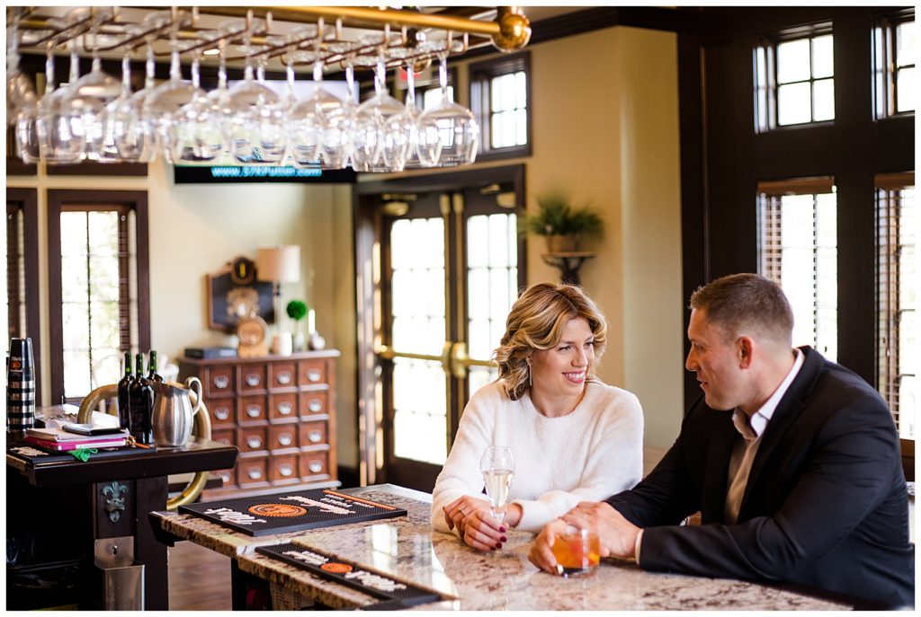 A photograph of an engaged couple sitting together, smiling and talking, in a classy club bar at Brookside Golf & Country Club by Columbus OH engagement photographer, Alayna Parker Photography